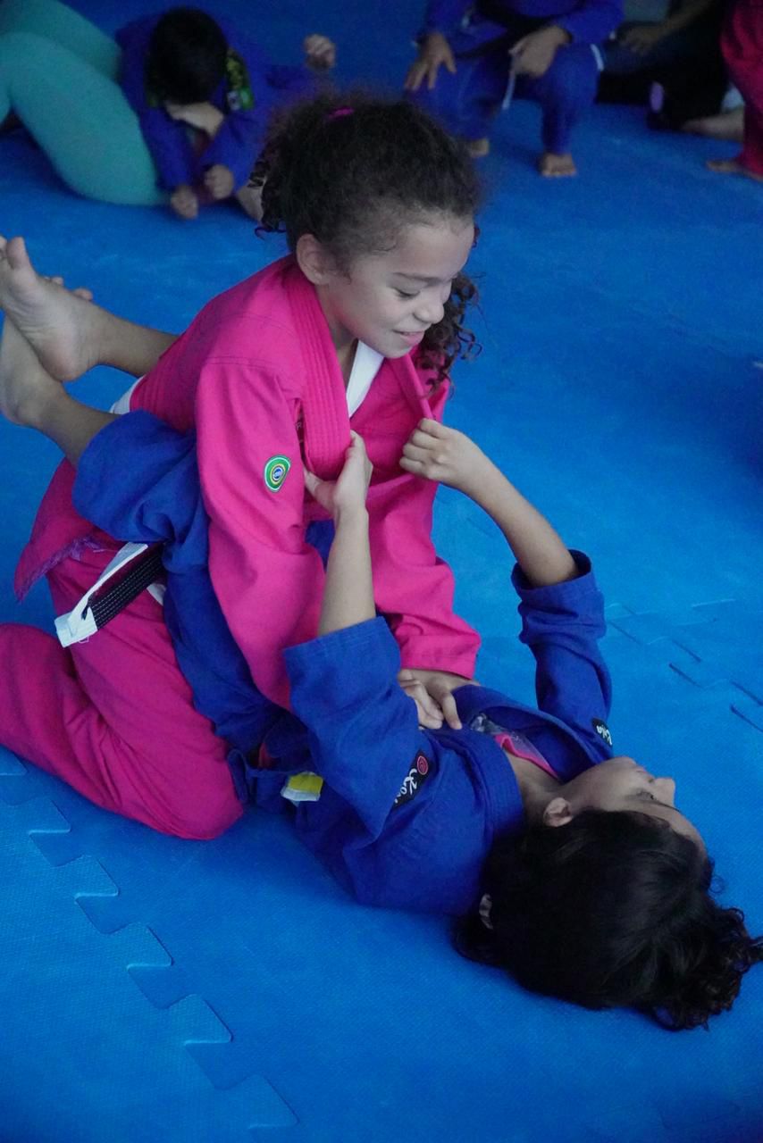 Aula infantil na GB Caruaru — menina de rosa passa guarda sobre colega de azul no tatame